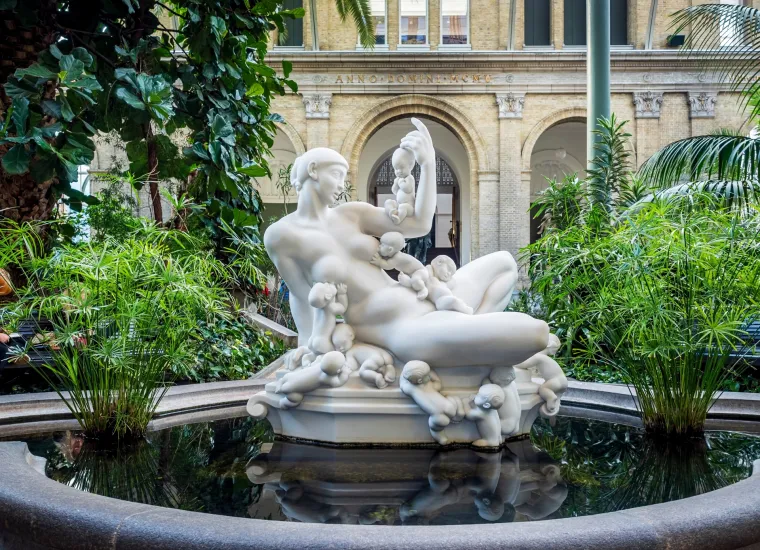 ZIMSKI VRT GLYPTOTEKET (DANSKA)- pravi je skriveni dragulj smje&scaron;ten u srcu Ny Carlsberg Glyptotek muzeja, koji je poznat po jedinstvenoj zbirci umjetnosti i skulptura. Ovaj zeleni raj s tropskim biljkama, palmama i egzotičnim cvijećem nalazi se pod staklenim krovom koji stvara atmosferu topline i mira, čak i tijekom hladnih zimskih dana.
Sredi&scaron;te vrta krasi fontana, čineći ambijent opu&scaron;tajućim, dok prozračni stakleni zidovi omogućuju prirodnom svjetlu da obasjava prostor. Zimski vrt Glyptoteket pruža savr&scaron;eno mjesto za predah posjetiteljima muzeja, koji nakon razgledavanja umjetničkih djela mogu uživati u prirodnoj ljepoti ovog prostora, evocirajući dojam mediteranskog vrta usred sjevera.