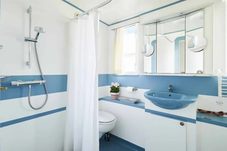 Osim kuhinje, ovaj luksuzni dom na vodi sadrži i tri elegantne kupaonice, uređene u svježim plavo-bijelim tonovima koji doprinose morskom ugođaju. Prva kupaonica opremljena je tuš kabinom s bijelim zavjesama koje stvaraju prozračan izgled, uz jednostavno ogledalo. 