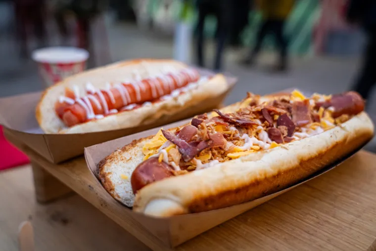 Rudolf&apos;s cheesy sausage naziv je ove zasitne kobasice koja je u mekanom pecivu poslužena sa sirom ementalerom, cheddar sirom, prženim lukom, hrskavom pancetom i GF umakom. 