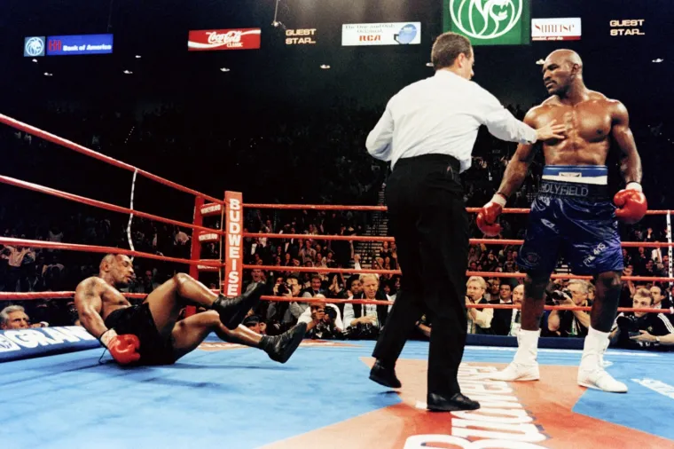 U njihovom meču prije toga održanom 9. studenog 1996., Holyfield je u borbi za tri teškaška pojasa tehničkim nokautom pobijedio Mike Tysona u Las Vegasu, na istom mjestu na kojem će manje od godinu dana kasnije imati meč u kojem će doći do spomenutog incidenta. Tyson u ovom meču nije znao što ga je snašlo. Sudac Mills Lane na ovoj fotografiji zaustavlja Evandera i prekida meč. Evander je bio izazivač. Boksački svijet ostao je zapanjen i šokiran načinom i dominacijom Evandera Holyfielda u toj borbi. 