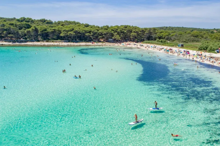3. Sakarun, Dugi Otok: Ova je plaža poznata po kristalno čistom moru i bijelom pijesku, što je rijetkost na hrvatskoj obali. Okružena je borovom šumom, koja pruža ugodan hlad, a plitko more idealno je za obitelji s djecom. Većina restorana i barova na plaži radi sezonski, pa je preporučljivo provjeriti radno vrijeme prije odlaska. Možete odsjesti u obližnjem hotelu Villa Nai 3.3 ili iznajmiti smještaj uz plažu. Sakarun se često naziva &quot;Karibima Jadrana&quot;.