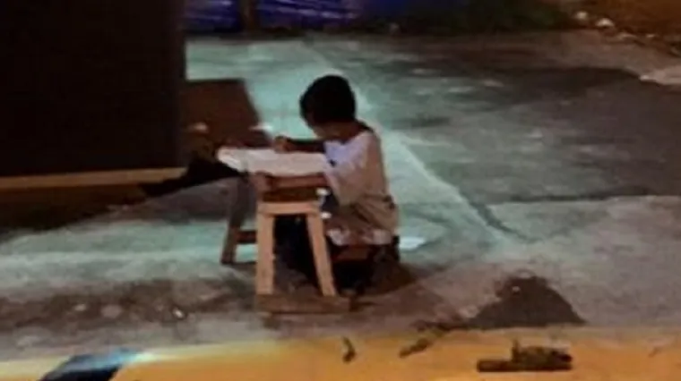 Maleni beskućnik piše zadaću na cesti uz svjetlo iz restorana